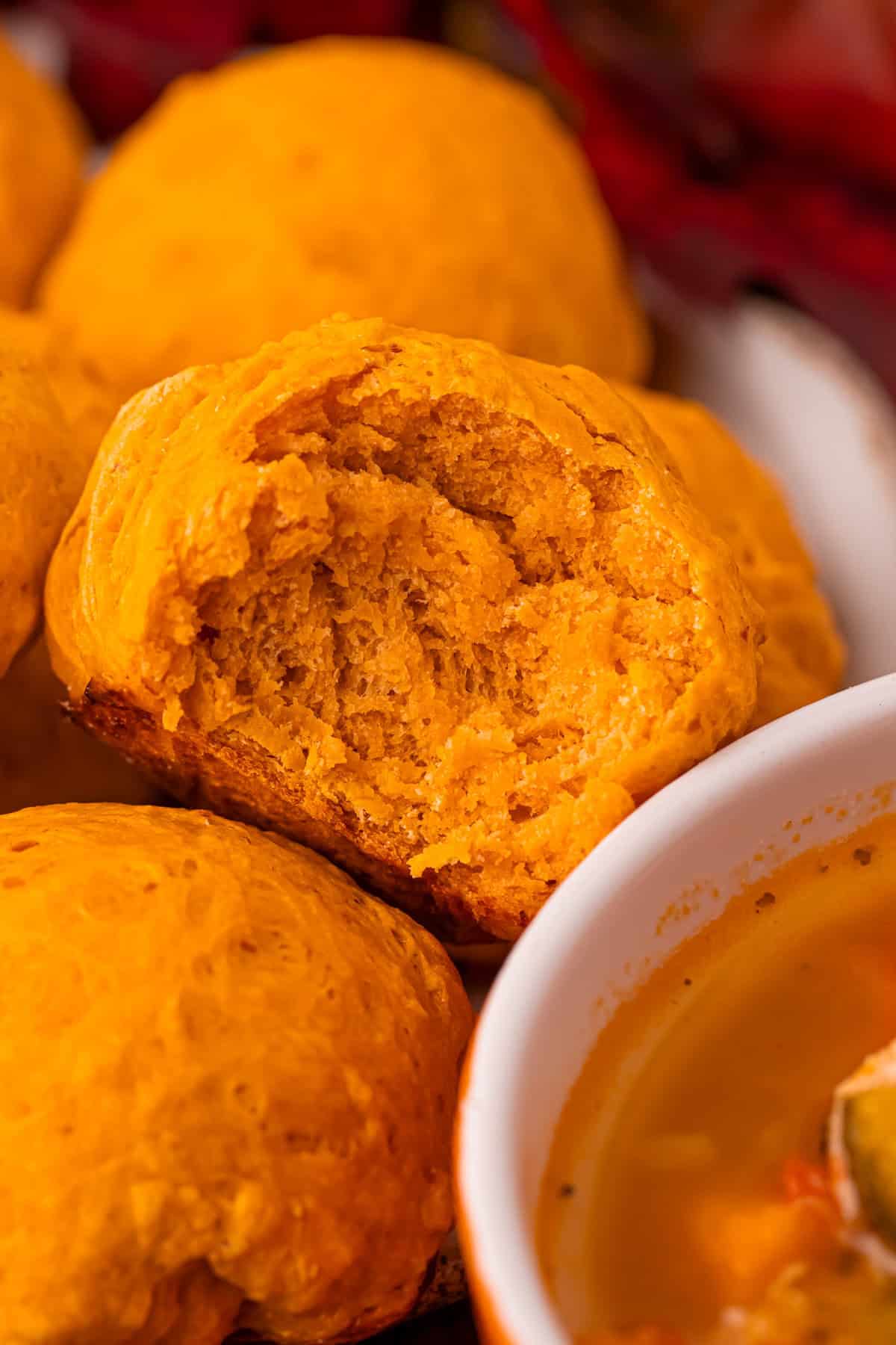 Close-up shot of a pumpkin bread roll with a bite revealing its soft, fluffy interior. Other rolls and part of a bowl of soup are visible in the background.