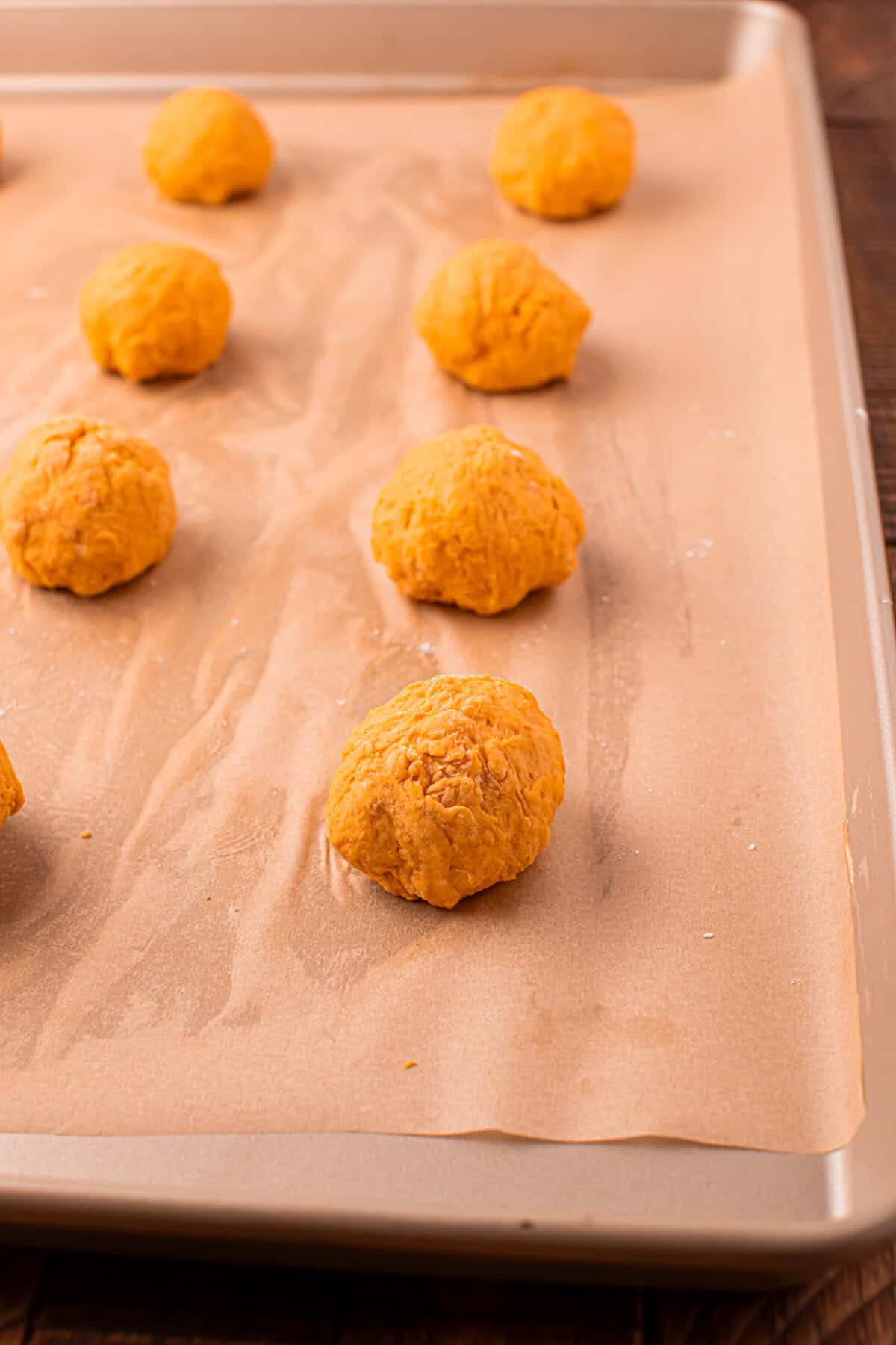 Unbaked pumpkin dough balls are spaced out on a parchment-lined baking sheet, ready for baking. The dough is light orange and slightly rough in texture.
