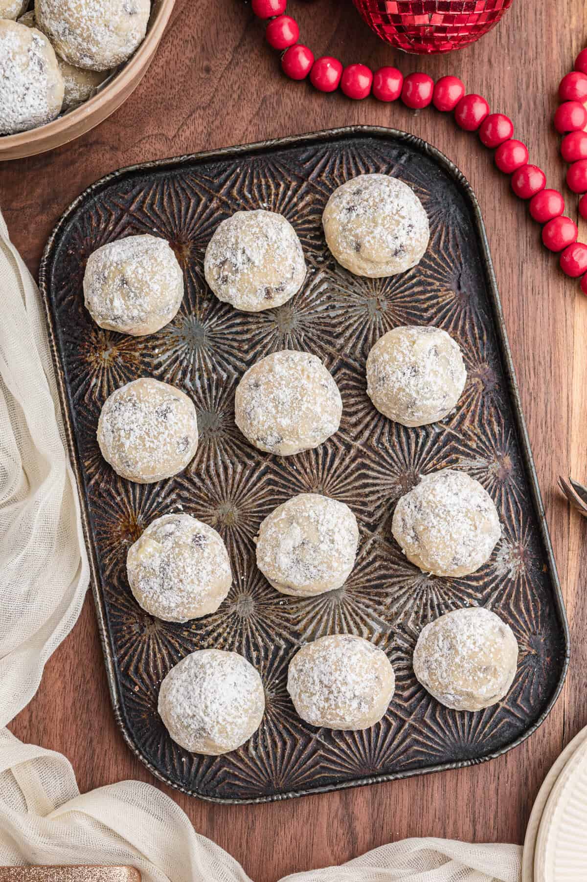 Snowball cookies with chocolate chips coated in confectioners sugar on a dark patterned tray arranged in neat rows.