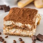 A tiramisu slice sits on a white plate surrounded by coffee beans, chocolate chunks, and ladyfingers.