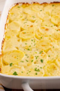 Close-up of creamy scalloped potatoes baked in a white rectangular dish. The potatoes are golden on top, with a light sprinkle of chopped parsley and browned edges along the pan.