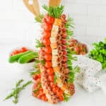 A tree-shaped appetizer display made of skewers of cherry tomatoes, cubed cheese, rolled salami slices, grapes, and rosemary sprigs, all arranged symmetrically around the form.