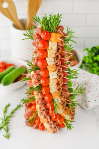 A charcuterie christmas tree appetizer with skewers forming a cone-shaped tree. Each skewer holds cherry tomatoes, cubes of cheese, rolled salami, and grapes, with sprigs of rosemary interspersed for garnish.