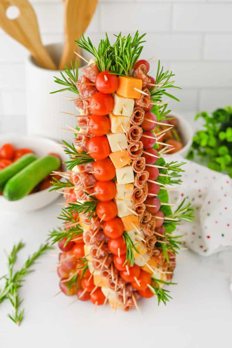 A charcuterie christmas tree appetizer with skewers forming a cone-shaped tree. Each skewer holds cherry tomatoes, cubes of cheese, rolled salami, and grapes, with sprigs of rosemary interspersed for garnish.