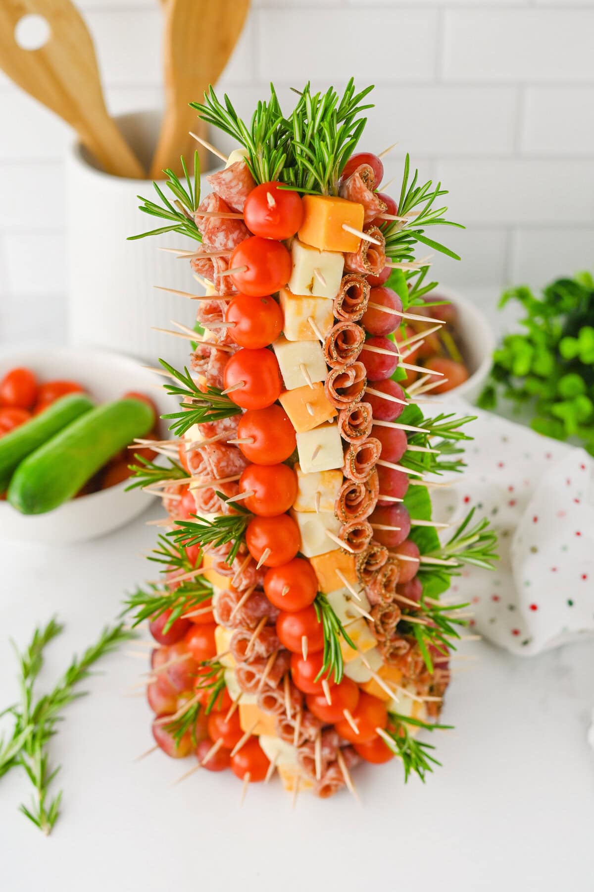 A charcuterie christmas tree appetizer with skewers forming a cone-shaped tree. Each skewer holds cherry tomatoes, cubes of cheese, rolled salami, and grapes, with sprigs of rosemary interspersed for garnish.