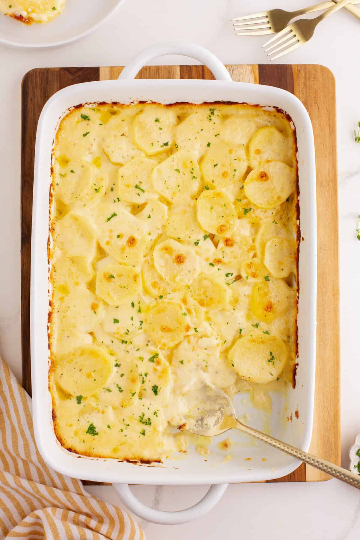 Top-down view of a white baking dish with scalloped potatoes, partially scooped out. The creamy potatoes are garnished with parsley, and the edges of the dish show golden browning. A serving spoon rests in the dish.