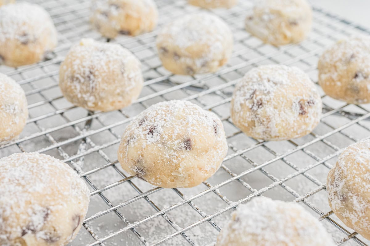 Several chocolate chip snowball cookies dusted with powdered sugar are arranged on a metal cooling rack. The cookies are round with a light golden color and specks of chocolate chips visible through the sugar coating.