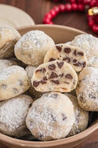 Chocolate chip snowball cookies in a bowl. The cookies are coated in powdered sugar, and a halved cookie on top shows the interior full of mini chocolate chips.