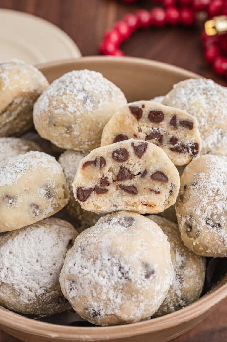 Chocolate chip snowball cookies in a bowl. The cookies are coated in powdered sugar, and a halved cookie on top shows the interior full of mini chocolate chips.