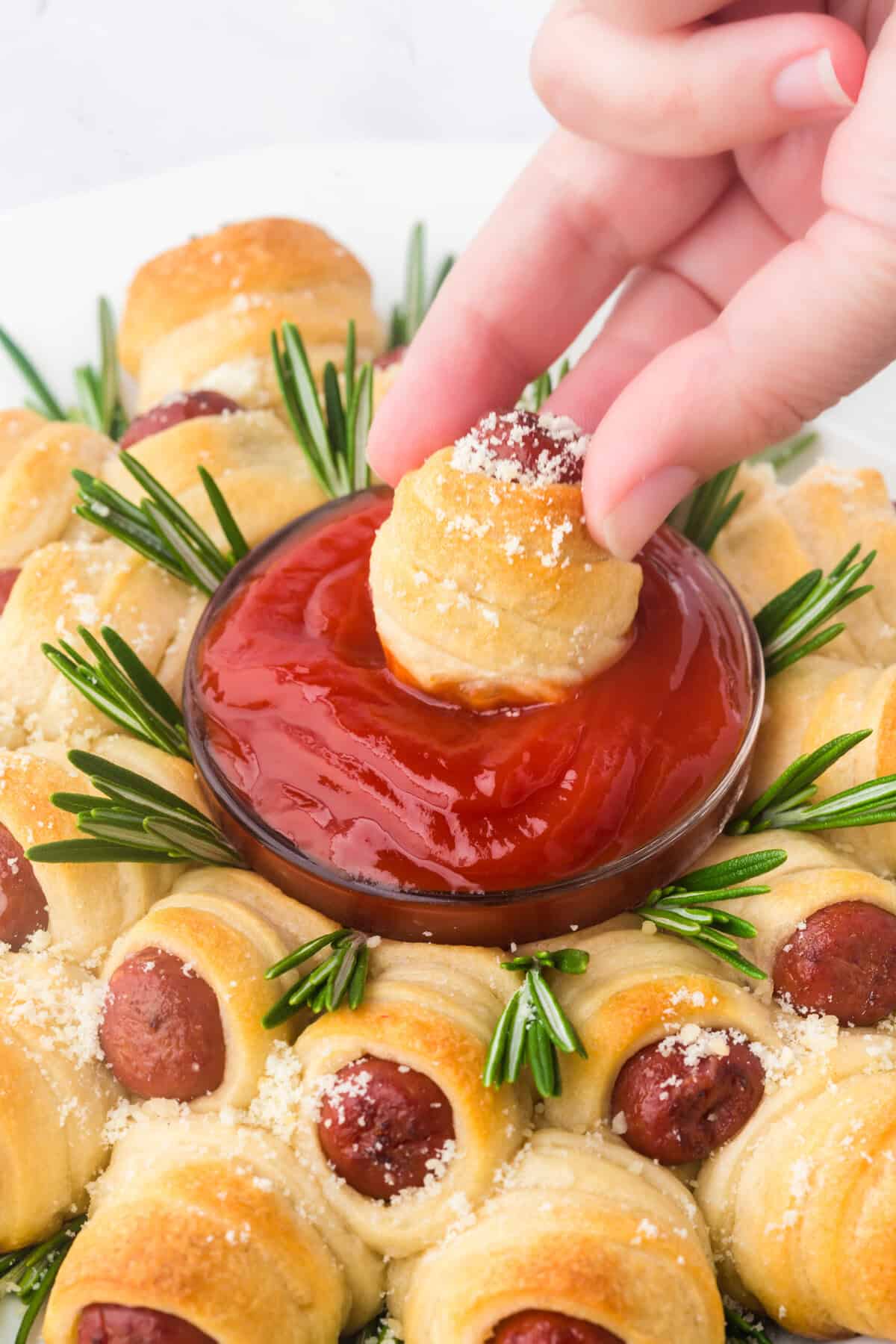 A hand dipping a crescent roll-wrapped sausage into a bowl of ketchup, surrounded by similar sausage rolls arranged in a ring with rosemary sprigs and grated cheese.