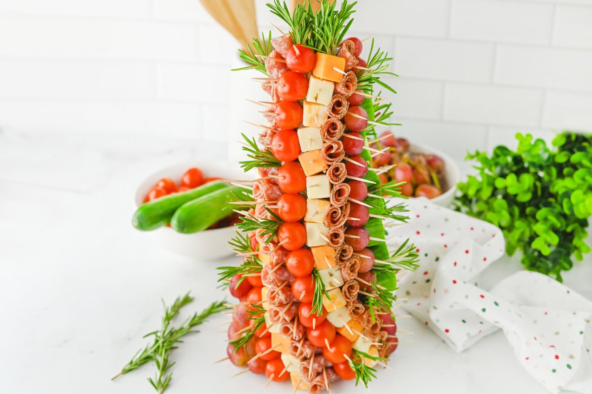 A festive Christmas appetizer tree made featuring cherry tomatoes, cubed cheeses, rolled salami, and grapes. The skewers are inserted into a foam cone, and rosemary sprigs are used to add greenery.