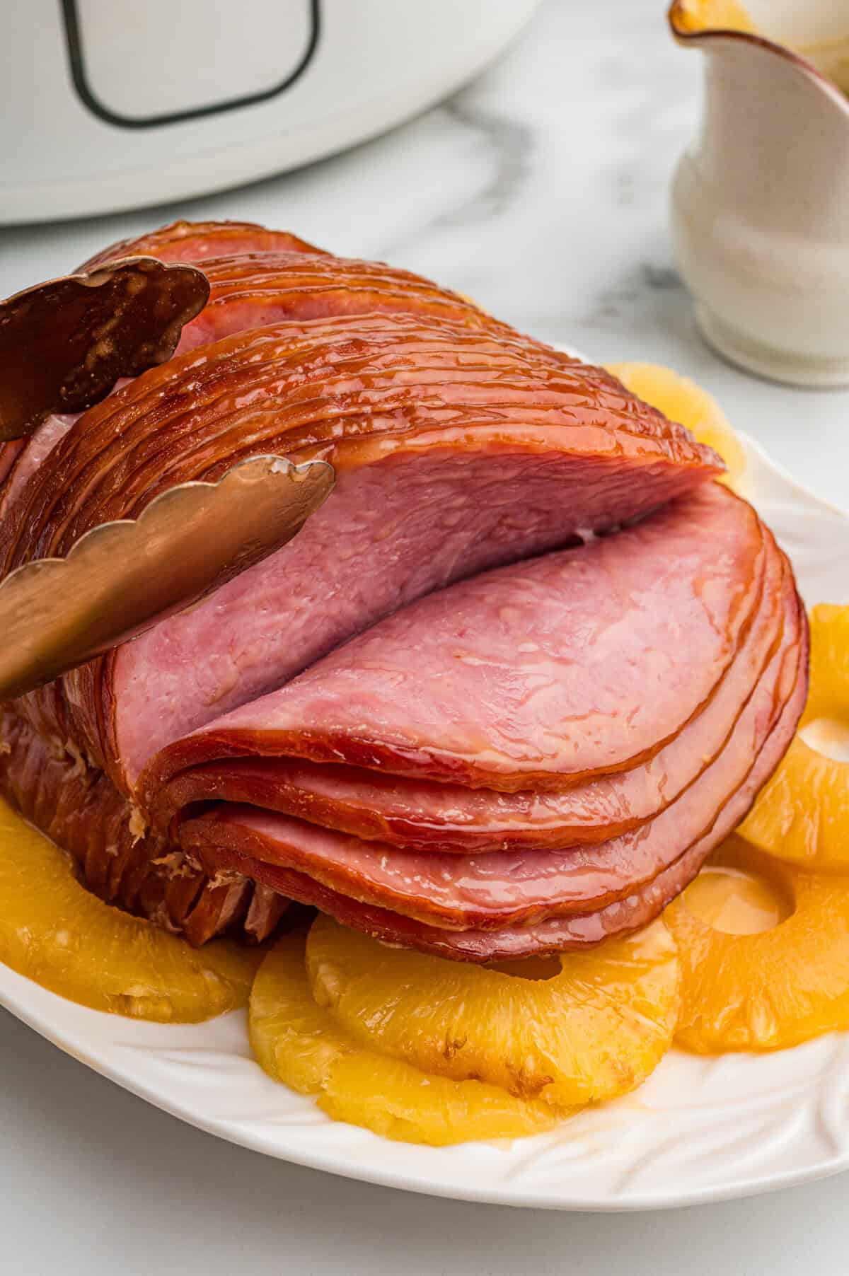 A platter of boneless spiral sliced ham surrounded by pineapple rings. Metal tongs are holding a slice of ham. The ham is glazed and slightly shiny. A crockpot is in the background.