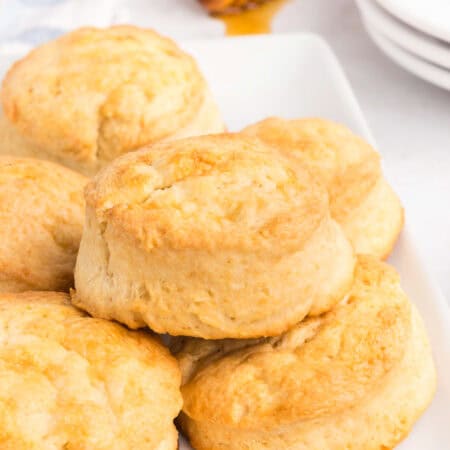 Overhead view of multiple golden brown biscuits piled on a white serving platter, with a honey dipper, bowl of honey, and stacked plates visible in the background.