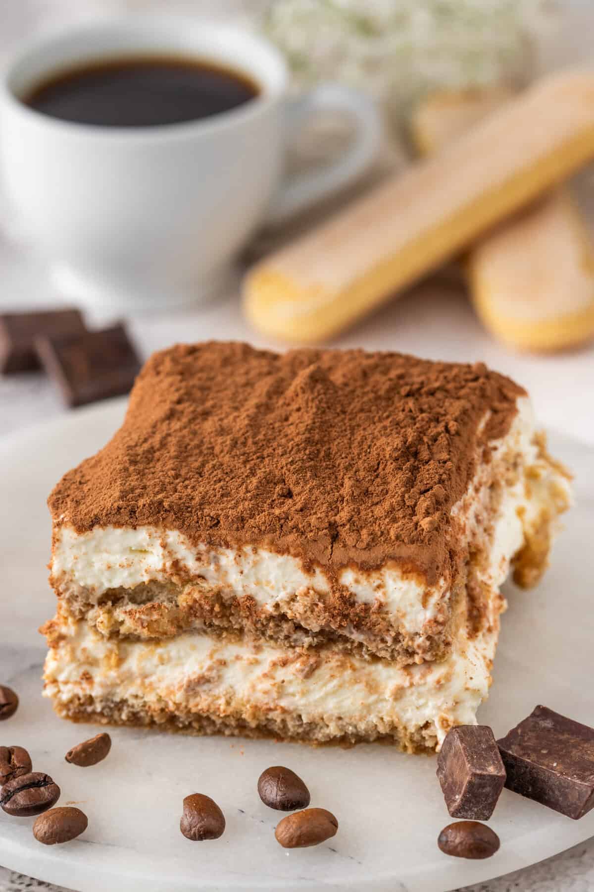 A tiramisu slice sits on a white plate surrounded by coffee beans and chocolate chunks. A white cup of coffee, ladyfinger cookies, and blurred flowers are in the background.