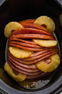 A glazed spiral sliced ham sits in a black slow cooker with pineapple rings arranged between the slices and around the edges.