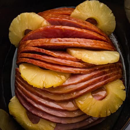 A glazed spiral sliced ham sits in a black slow cooker with pineapple rings arranged between the slices and around the edges.