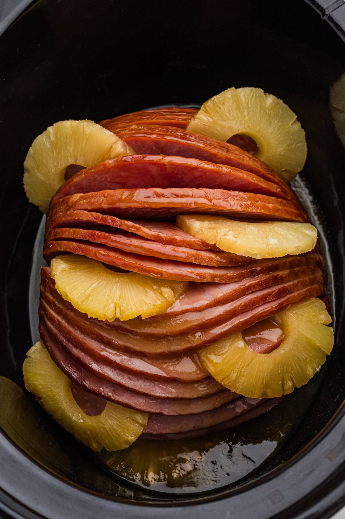 A glazed spiral sliced ham sits in a black slow cooker with pineapple rings arranged between the slices and around the edges.