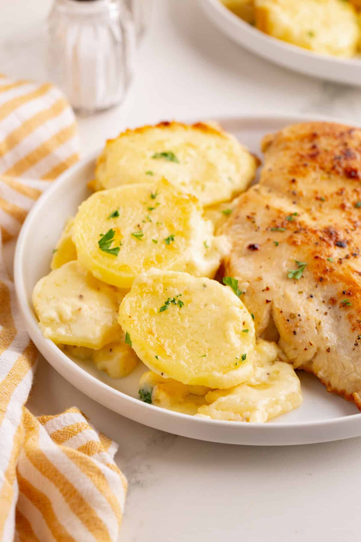 Homemade scalloped potatoes and a seasoned chicken breast on a white plate. The potatoes are creamy and topped with chopped parsley. 
