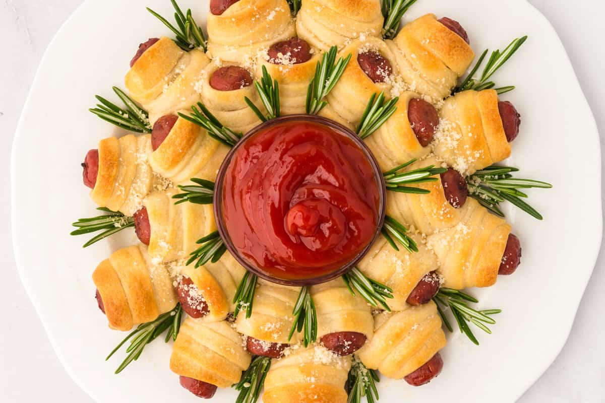 A wreath of crescent-wrapped mini hot dogs garnished with rosemary, surrounding a central glass bowl filled with ketchup, all on a light surface.