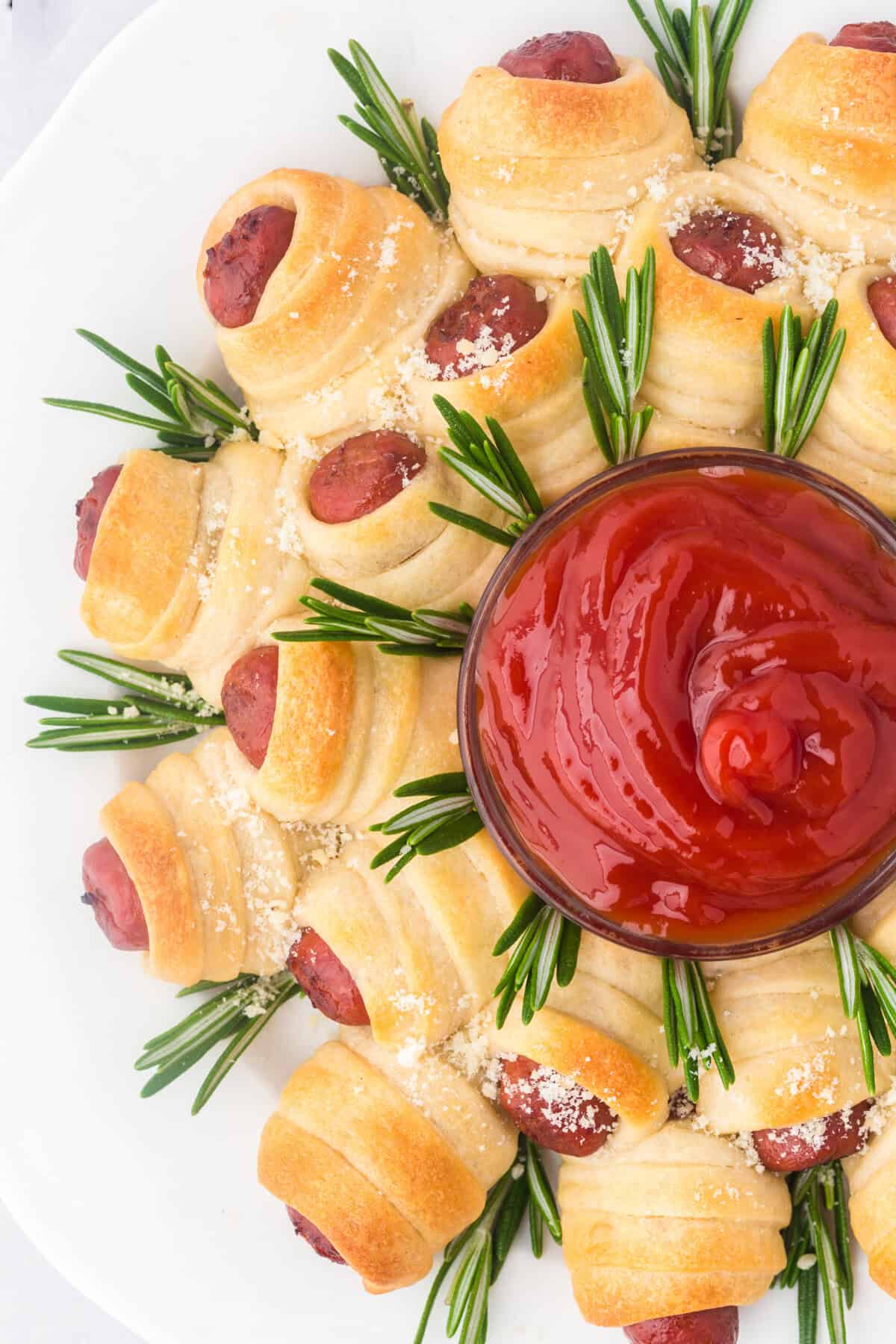 Overhead view of golden crescent roll-wrapped sausages arranged in a circular wreath pattern on a white plate, garnished with rosemary sprigs and grated cheese, with a bowl of ketchup in the center.