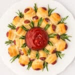 A pigs in a blanket wreath christmas appetizer made of golden crescent-wrapped lil smokies with rosemary and grated cheese, circling a bowl of ketchup on a white plate.