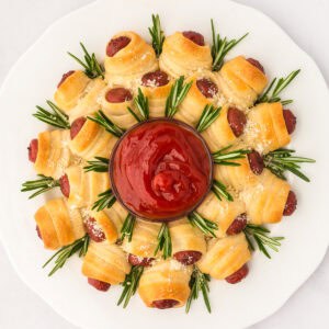 A pigs in a blanket wreath christmas appetizer made of golden crescent-wrapped lil smokies with rosemary and grated cheese, circling a bowl of ketchup on a white plate.