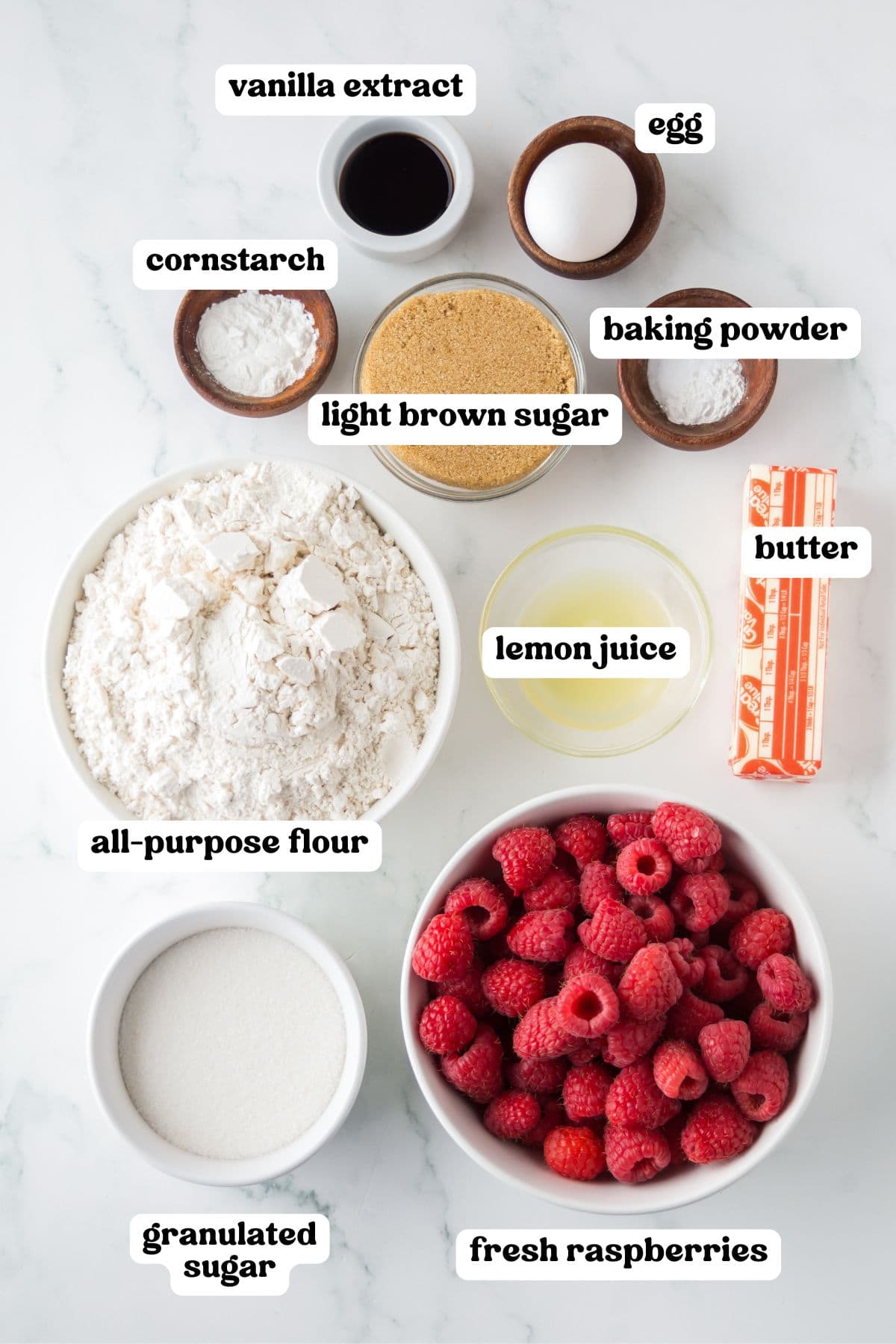 Flat lay of labeled ingredients for raspberry crumb bars arranged on a white surface, including fresh raspberries, all-purpose flour, granulated sugar, light brown sugar, lemon juice, butter, egg, baking powder, cornstarch, and vanilla extract.
