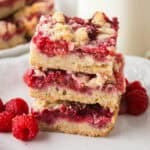 Stack of three raspberry crumble shortbread bars on a white plate with fresh raspberries.