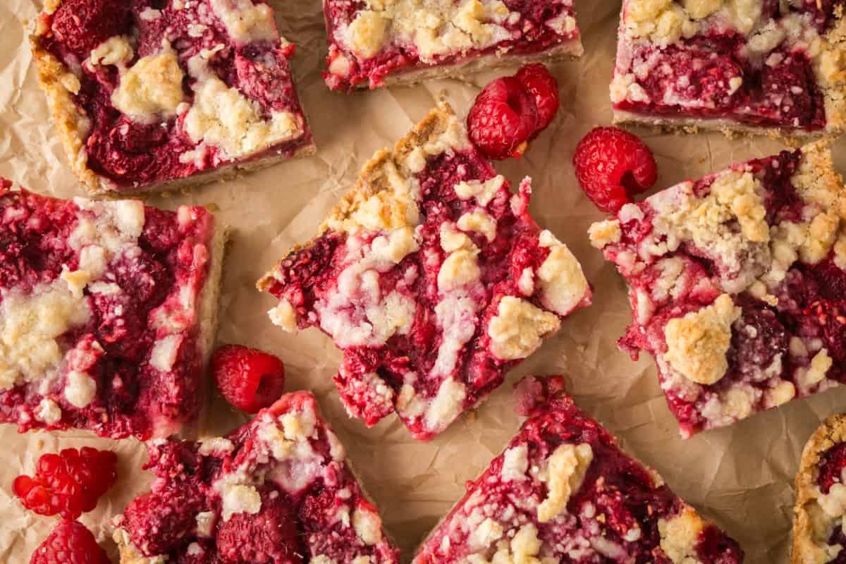 Overhead view of multiple raspberry crumb bars arranged on crumpled parchment paper, with some fresh raspberries scattered among the bars. Each bar has a golden crust, vibrant red raspberry filling, and a crumbly, pale shortbread crumb topping.