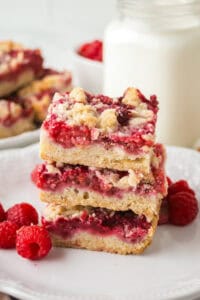stack of three raspberry crumble bars on a white plate, next to a glass jar of milk. The bars have golden crusts, thick raspberry layers, and crumbly toppings. Fresh raspberries are scattered around the plate.