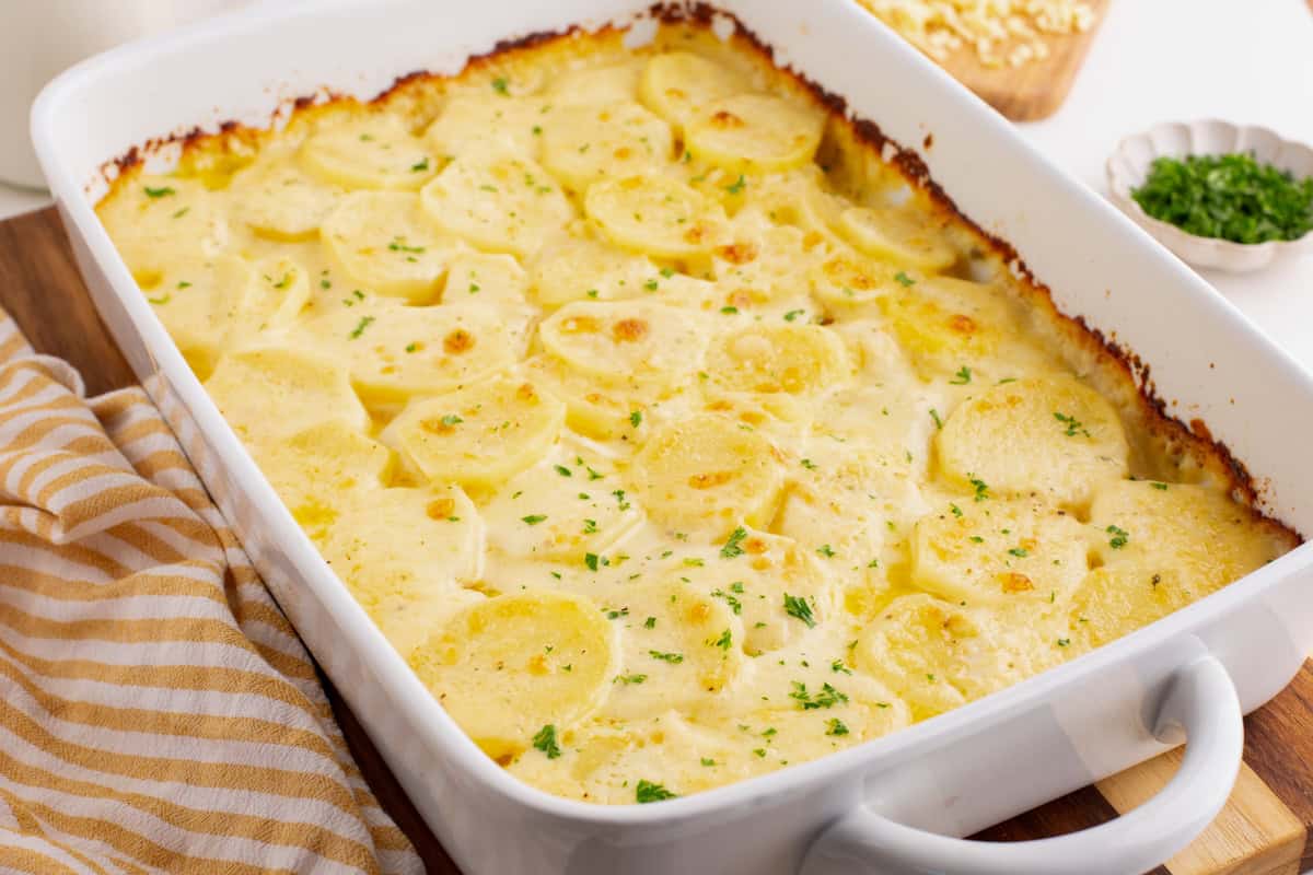 Dish filled with baked scalloped potatoes, showing lightly browned edges and parsley garnish. Chopped herbs and cheese are in the background.