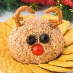 A reindeer cheese ball coated in chopped nuts, with black olives for eyes, a cherry tomato for a nose, and pretzel twists shaped into antlers. The cheese ball is on a plate surrounded by crackers, with holiday decorations in the background.