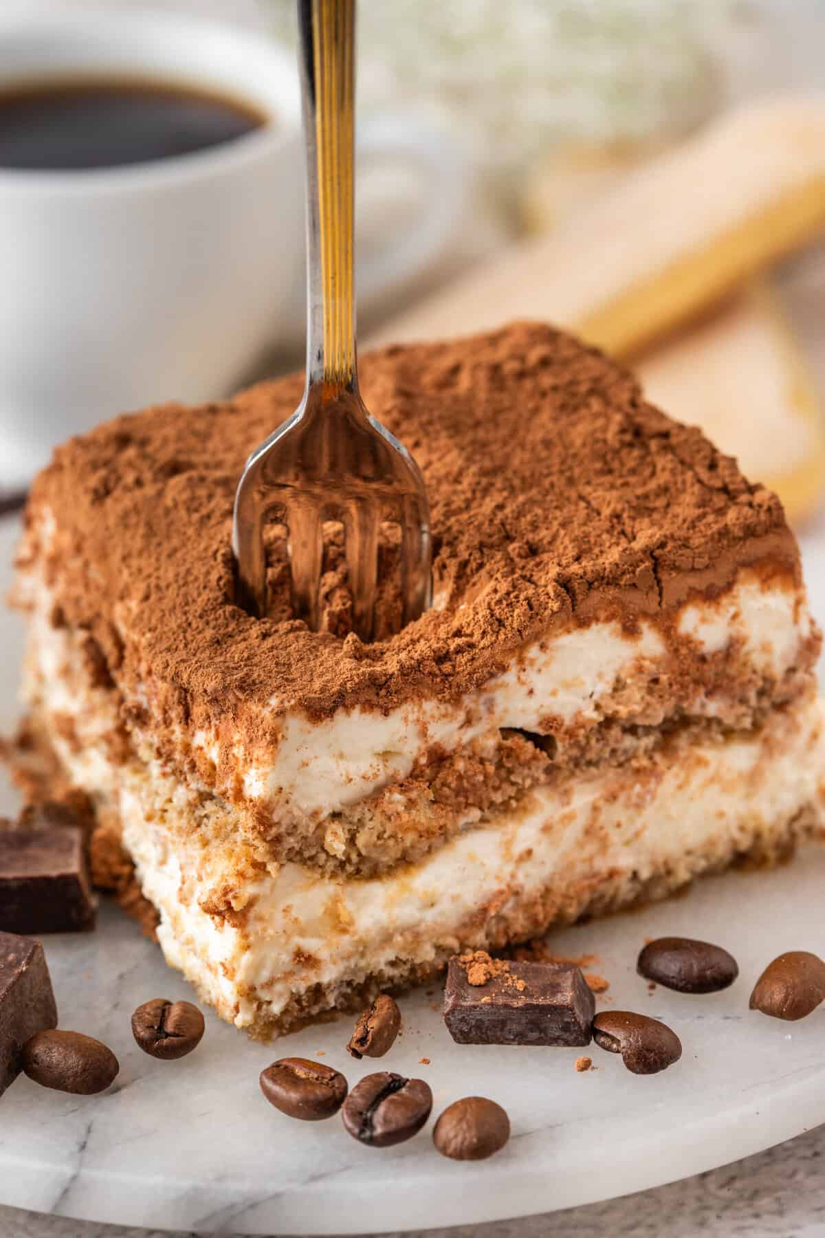 A fork is pressed into the top of a cocoa-dusted tiramisu dessert, revealing layers of cream and soaked ladyfingers. Coffee beans and chocolate chunks are scattered on the marble plate below.