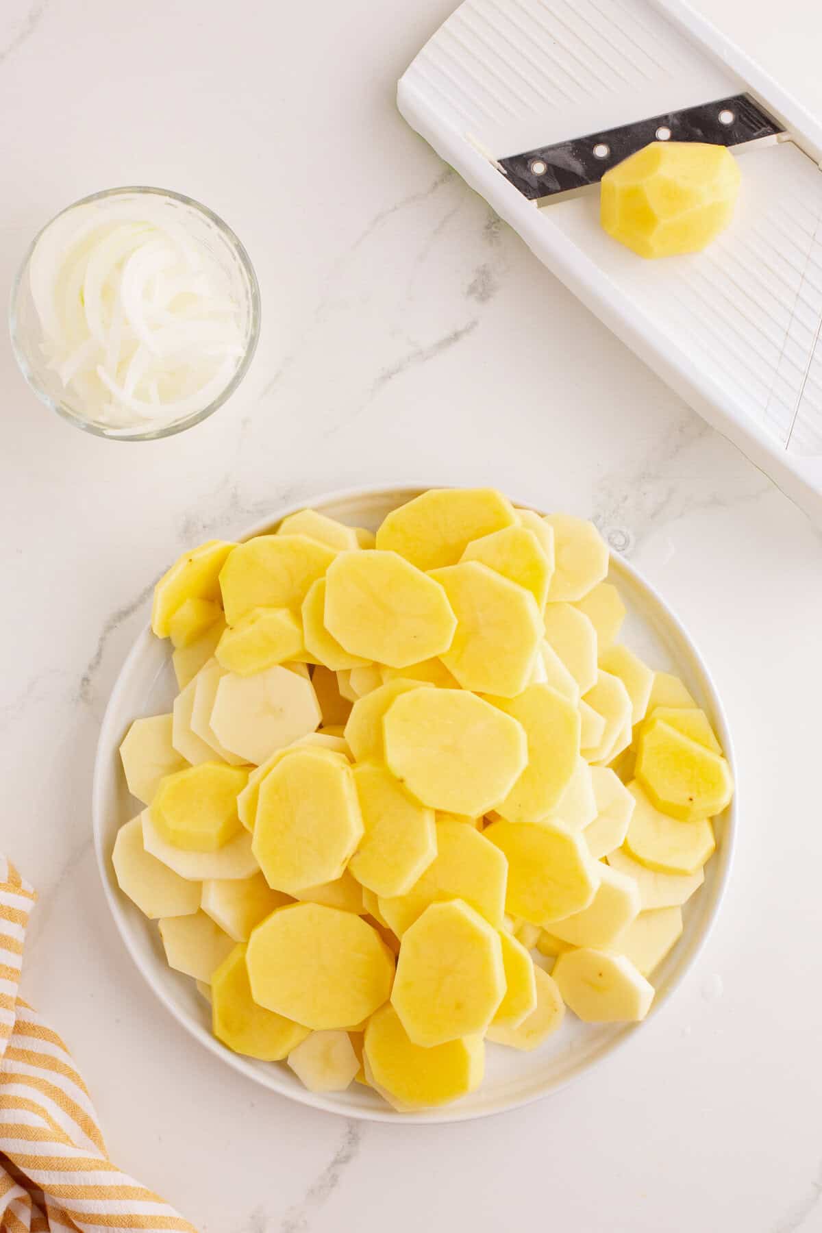 Plate of sliced potatoes and bowl of onions next to a mandoline slicer.