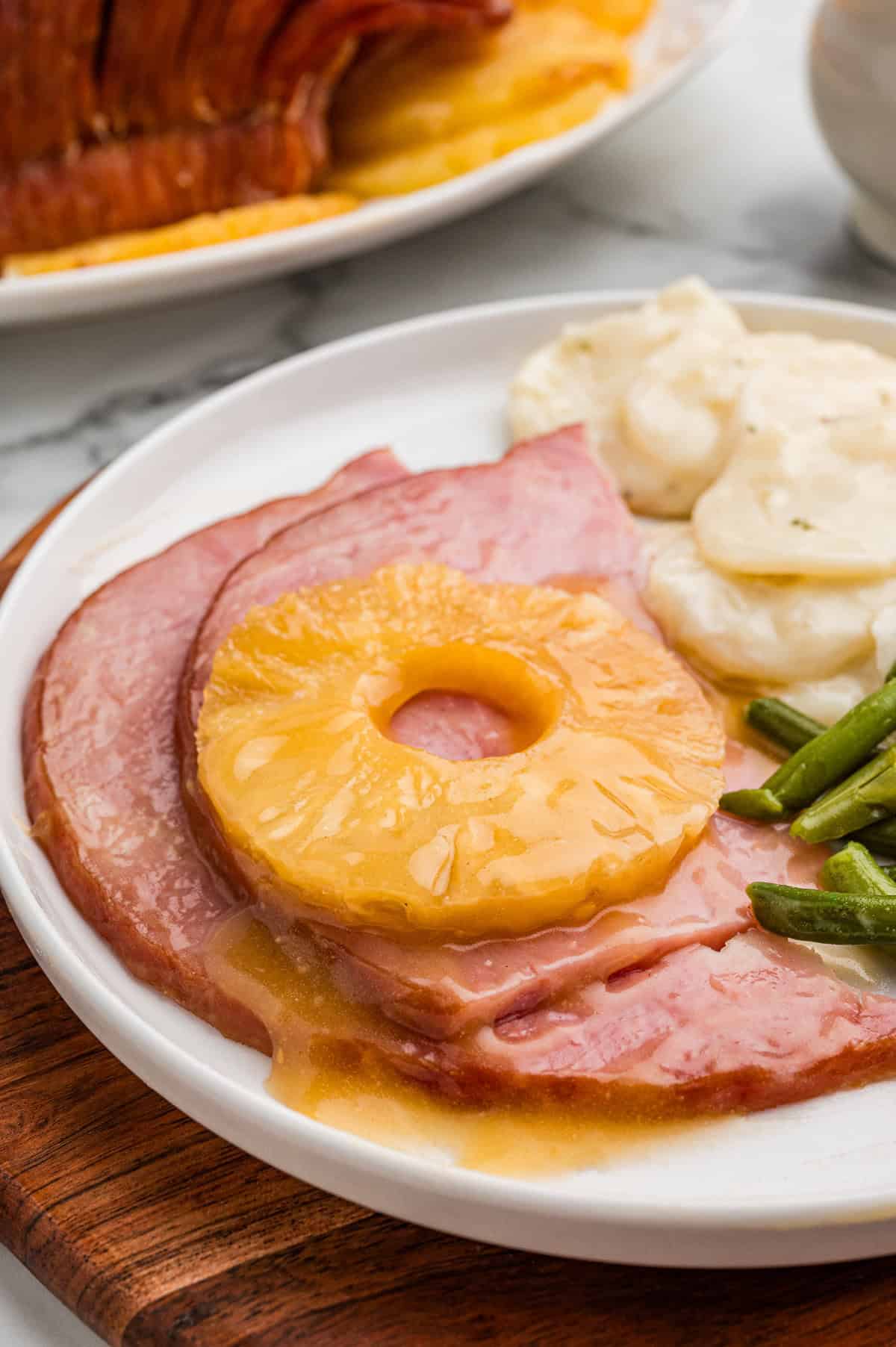 Close-up of sliced boneless spiral ham topped with a ring of pineapple on a white plate. The plate also contains creamy scalloped potatoes and green beans.