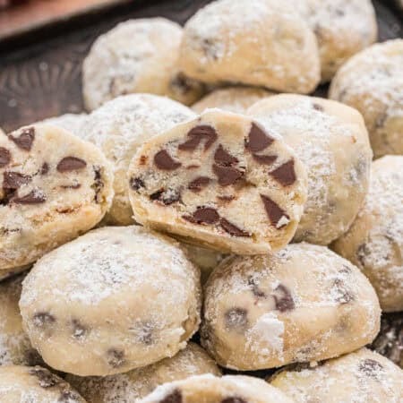 Mini chocolate chip snowball cookies stacked on a tray, with one cut open to show the chocolate chips inside. The cookies are coated in powdered sugar.