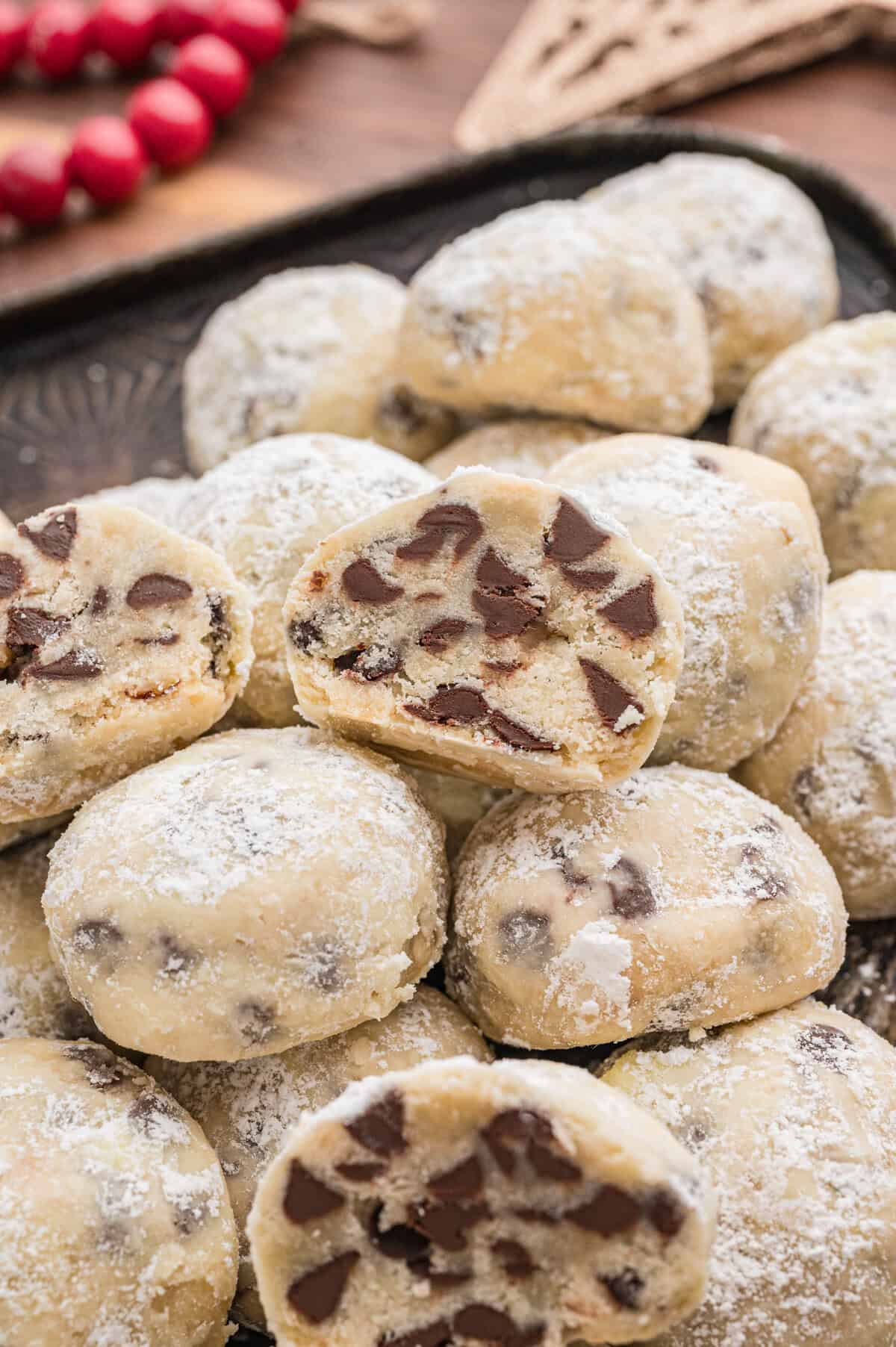 Close-up image of chocolate chip snowball cookies, with one cut open to show the soft, crumbly inside filled with chocolate chips. The cookies are coated in powdered sugar.