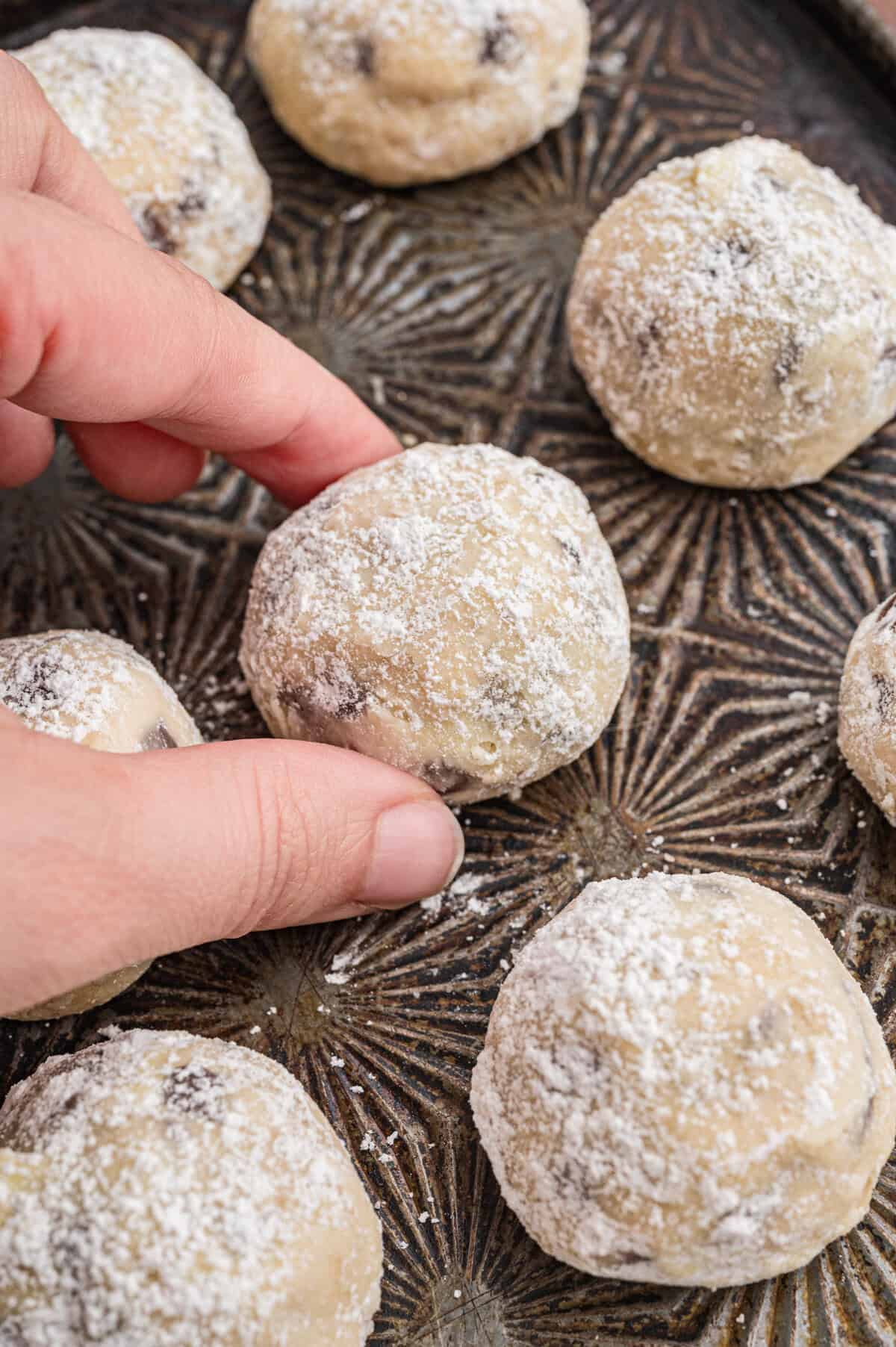 Close-up of a hand picking up a chocolate chip snowball cookie from a dark, patterned tray. The cookie is round, golden, and covered in powdered sugar. Several more cookies are visible on the tray.