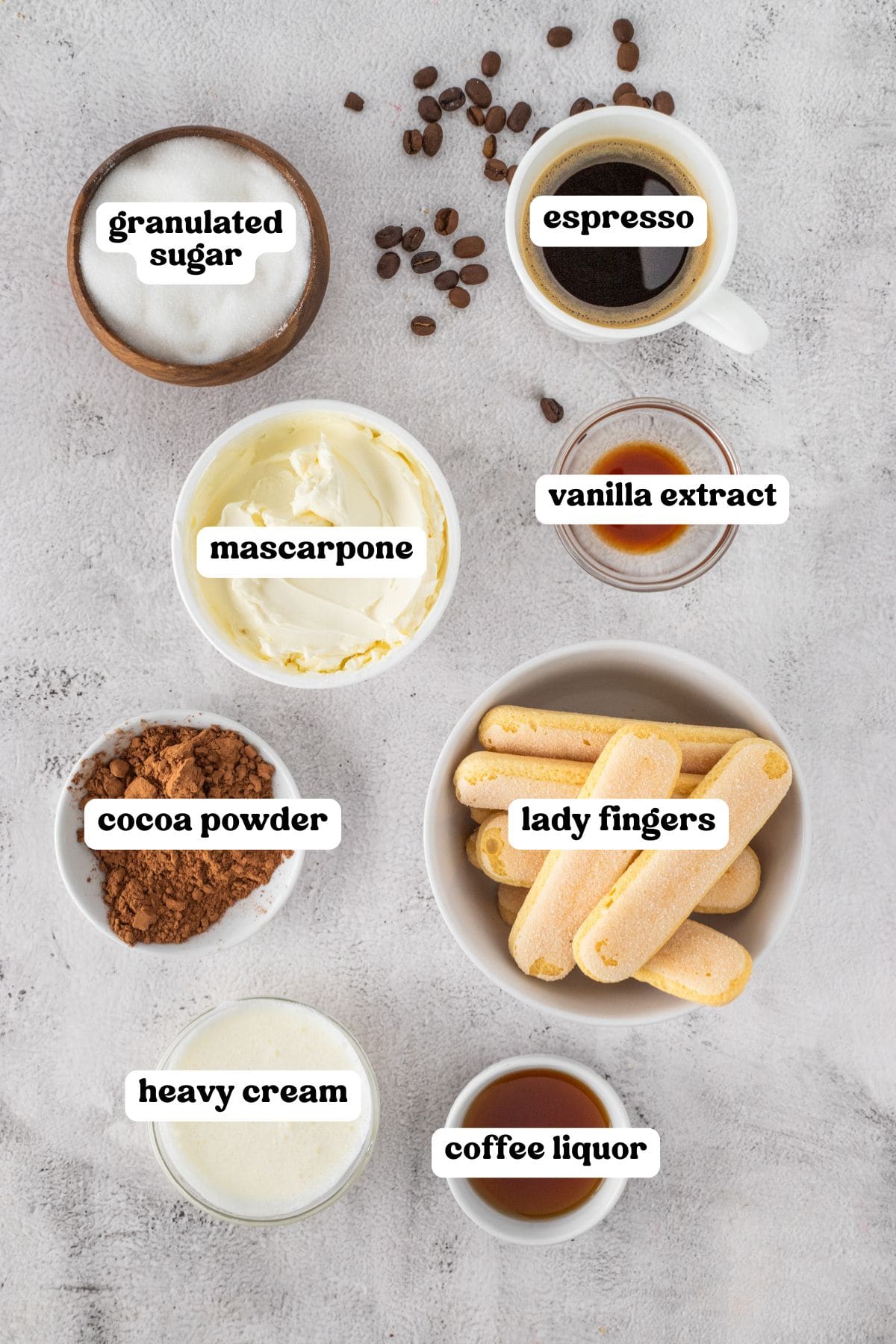 Labeled ingredients for tiramisu arranged on a white surface. Items include granulated sugar, espresso, mascarpone, vanilla extract, cocoa powder, lady fingers, heavy cream, and coffee liquor. Scattered coffee beans are visible around the items.