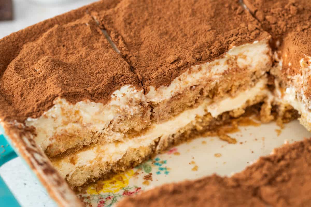 A rectangular dish of tiramisu with 2 pieces removed, revealing creamy layers of mascarpone and soaked ladyfingers beneath a thick layer of cocoa powder.