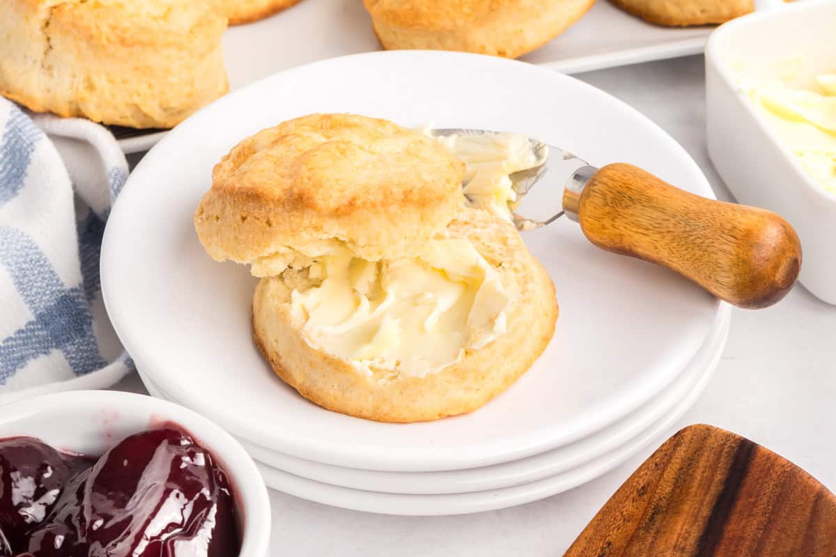 Close-up of a biscuit cut in half with softened butter spread on the bottom half using a wooden-handled knife, placed on a stack of white plates next to a bowl of jam.