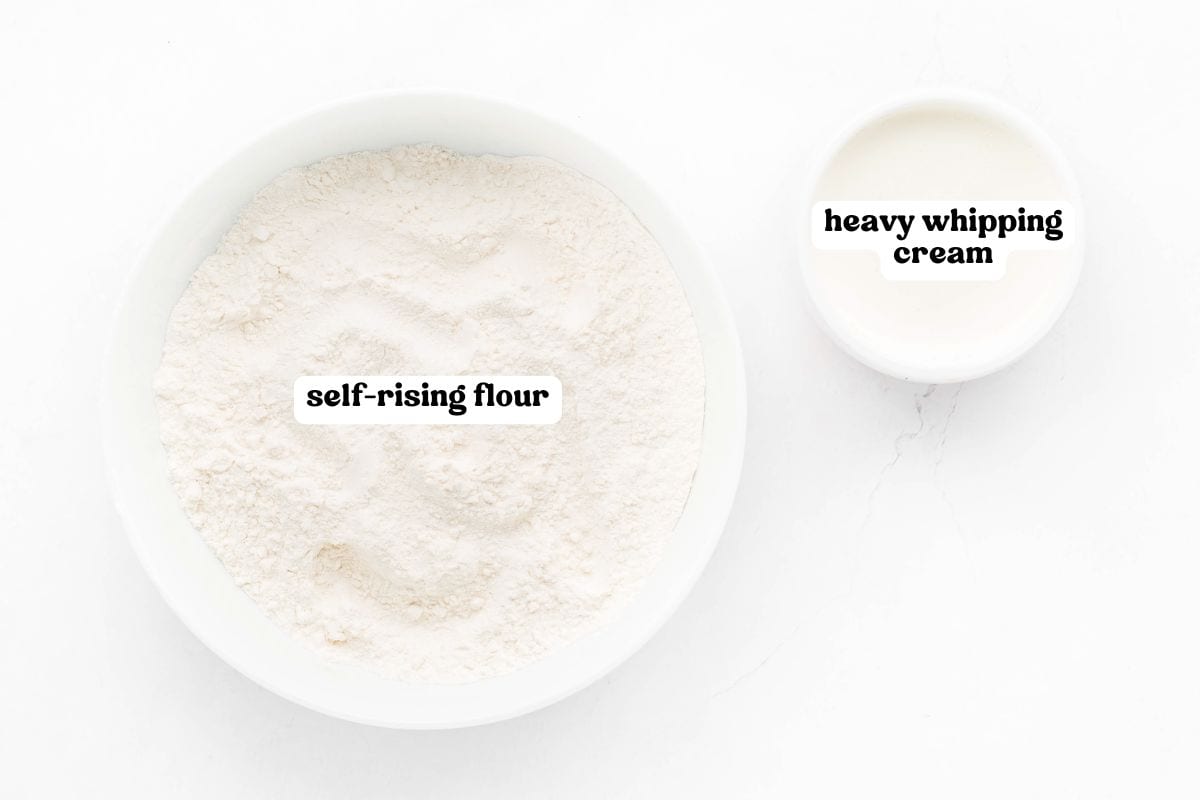 Overhead image of two white bowls on a light surface—one large bowl filled with self-rising flour and one smaller bowl with heavy whipping cream. Both bowls are clearly labeled with bold black text.