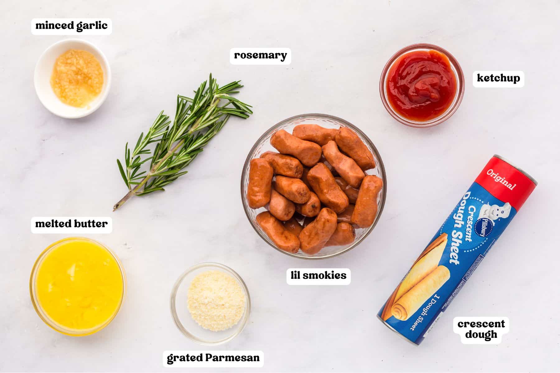 Overhead view of labeled ingredients, including minced garlic, rosemary sprig, ketchup, melted butter, grated Parmesan, a bowl of mini sausages labeled “lil smokies,” and a can of Pillsbury Crescent Dough Sheet.