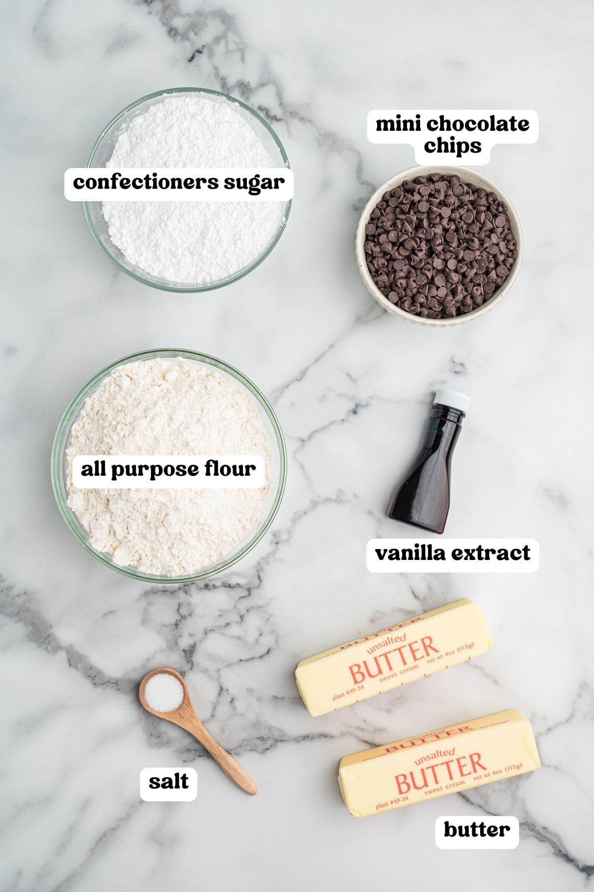 Overhead image of labeled baking ingredients including a bowl of confectioners sugar, a bowl of mini chocolate chips, a bowl of all-purpose flour, a bottle of vanilla extract, a spoonful of salt, and two sticks of unsalted butter.