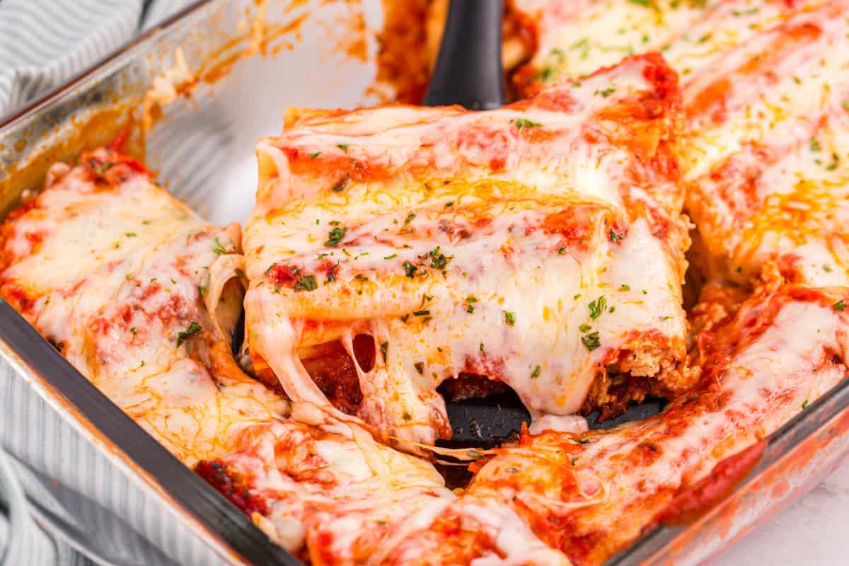 A spatula lifting a serving of baked cheese manicotti from the dish, with melted cheese stretching and filling visible inside the pasta tubes.