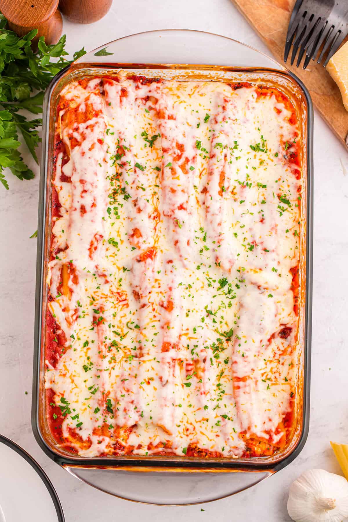 Overhead shot of a glass baking dish filled with neatly arranged manicotti covered in melted mozzarella and tomato sauce, garnished with chopped parsley.