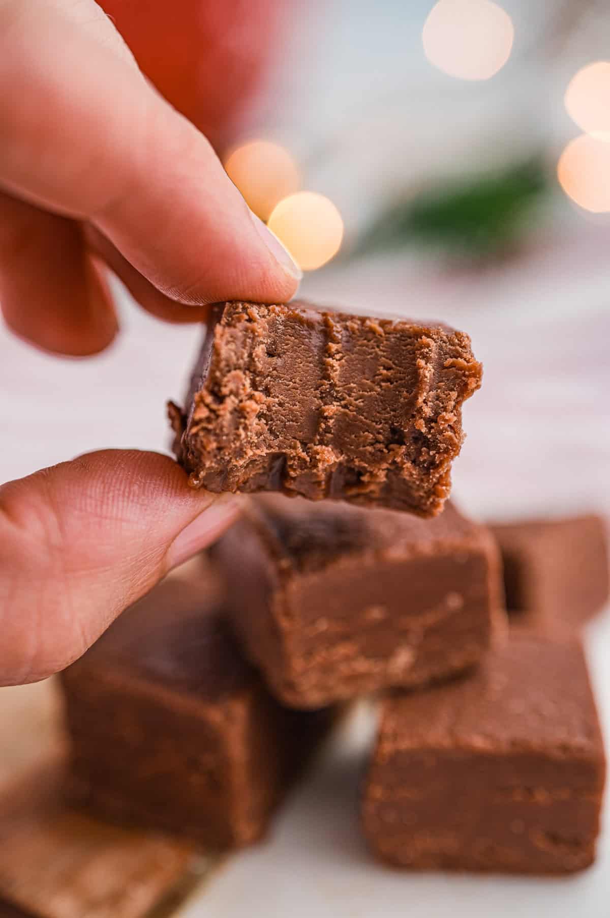 A close-up of a hand holding a square piece of chocolate fudge with a bite taken out, revealing the smooth, creamy interior.