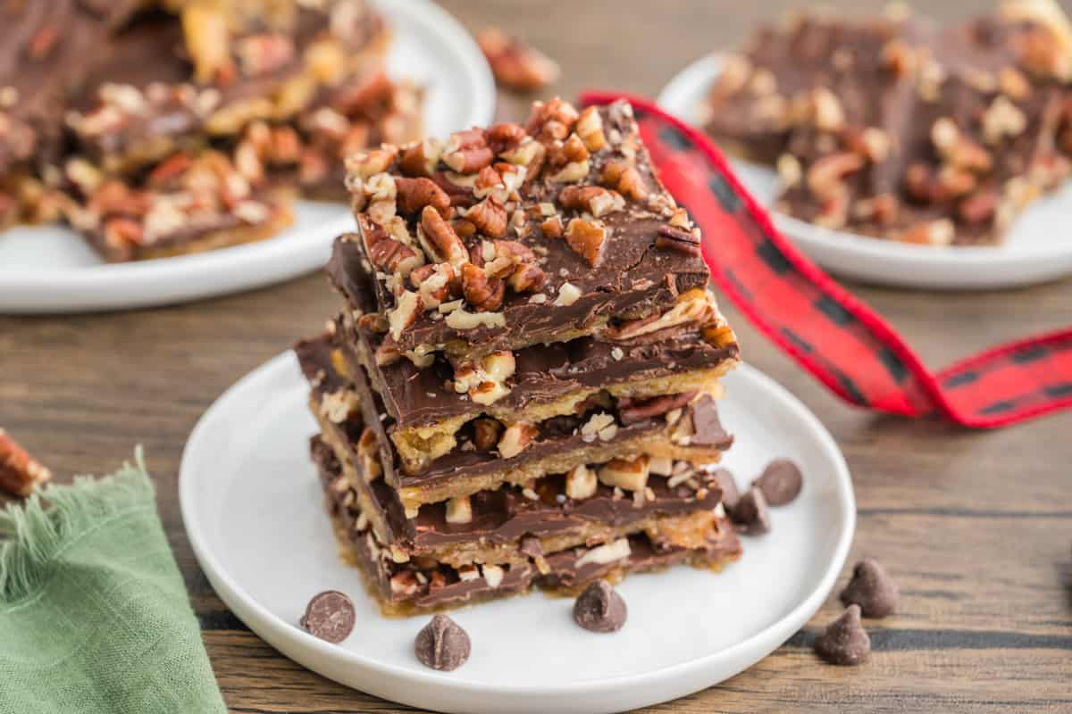 A stack of saltine cracker toffee topped with chopped pecans and layered with chocolate sits on a white plate. Chocolate chips are scattered around the plate, with blurred toffee pieces in the background.