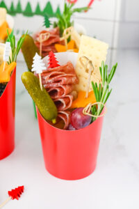 Christmas charcuterie cups - red paper cup filled with charcuterie ingredients: folded salami skewers, cheddar cheese stars, a gherkin, a grape, a cracker, and rosemary.