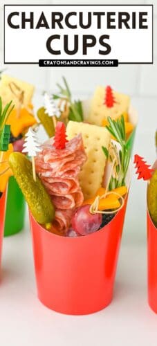 A red paper cup filled with salami, a gherkin, cheddar cheese star, grape, cracker, and rosemary. Topped with red and white tree picks. Text reads "CHARCUTERIE CUPS" and "crayonsandcravings.com."